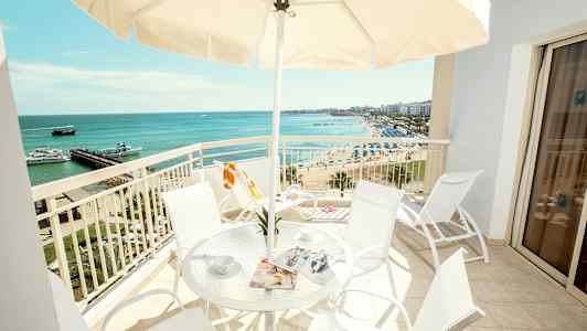 En balkong med bord og stoler, utsikt over stranden og havet. (AI-generert tekst)