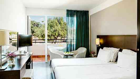 Hotellrom med to senger, et skrivebord og et lite bord med stoler foran en glassdør. (AI-generert tekst)