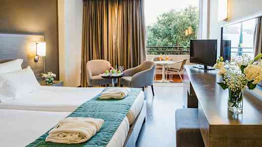 Et hotellrom med dobbeltseng, sittegruppe med to stoler og bord, og en balkong med utsikt. (AI-generert tekst)