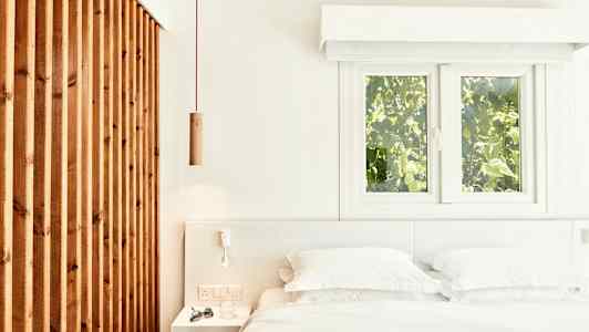 Lys og luftig soverominteriør med en seng, hvite sengeklær og en trepanel samt vinduer med utsikt til grøntområder. (AI-generert tekst)