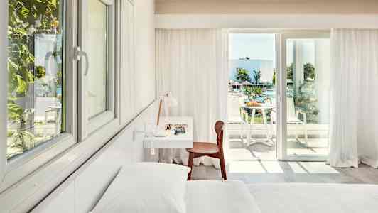 Interiør av et lyst hotellrom med seng, skrivebord og stol, samt en balkong med utsikt over et basseng og grøntområder. (AI-generert tekst)