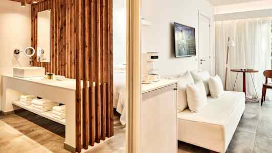 Et hotellrom med åpen planløsning, inkludert en TV, en sofa og et område med servant og badeartikler. (AI-generert tekst)