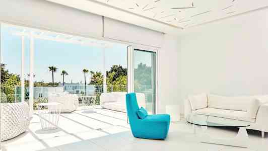 Moderne stue med store vinduer og utsikt mot en solrik terrasse med palmer. (AI-generert tekst)