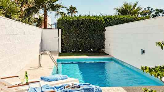 Et mindre basseng omgitt av hvite vegger og grønn vegetasjon, med to solsenger nær kanten. (AI-generert tekst)