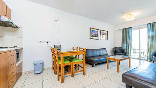 Et oppholdsrom med møbler inkludert et bord med stoler, en sofa og en lenestol. Et kjøkken er synlig i hjørnet og en balkong med dør i bakgrunnen. (AI-generert tekst)