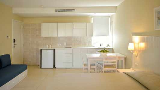 En lys og moderne studio med kjøkkenområde bestående av hvite skap, et lite bord med stoler og et sittemøbel mot en beige vegg. (AI-generert tekst)