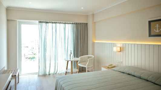 Bilde av et lyst hotellrom med en seng, et bord og en stol nær et vindu med lyse gardiner. (AI-generert tekst)