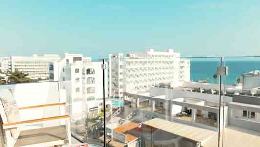 Utsikt fra en balkong med stoler og et bord, med hotellbygninger og havet i bakgrunnen. (AI-generert tekst)