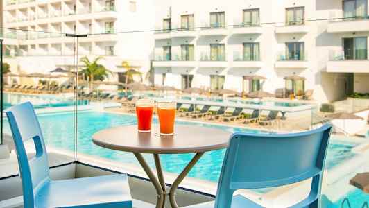 To fargerike drikker står på et bord ved siden av to stoler, med et basseng og hotell i bakgrunnen. (AI-generert tekst)