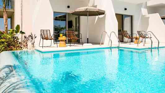 Et utendørs basseng foran en bygning med bord og stoler i bakgrunnen. (AI-generert tekst)