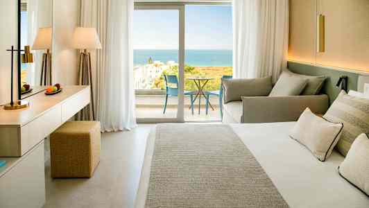 Et hotellrom med en dobbeltseng, sittegruppe og en balkong med utsikt mot havet. (AI-generert tekst)