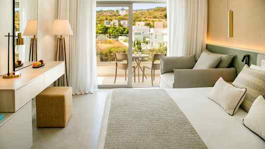 Bilde av et hotellrom med seng, sofa og skrivebord, samt en balkong med utsikt over et boligområde. (AI-generert tekst)