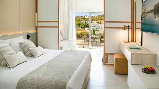 Et lyst hotellrom med en stor seng, en arbeidsplass og en balkong med utsikt over grøntarealer og bygninger. (AI-generert tekst)