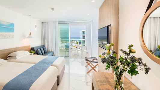 Et hotellrom med to senger, en sofa, et skrivebord med blomster, stort speil, og en balkong med utsikt mot havet. (AI-generert tekst)