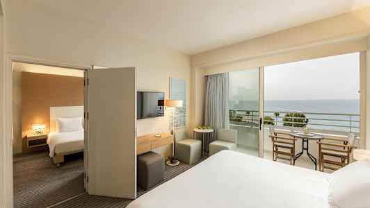 Et hotellrom med en seng, sitteplasser og et bord, og en balkong med utsikt over havet. (AI-generert tekst)