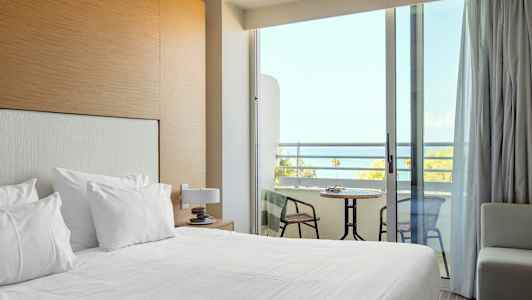 Et hotellrom med oppredd seng og en balkong med utsikt over havet. (AI-generert tekst)