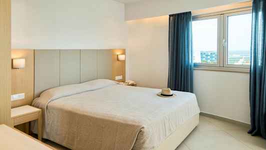 Et hotellrom med en stor seng, to nattbord, to lamper, et vindu med blå gardiner og en hatt på sengen. (AI-generert tekst)