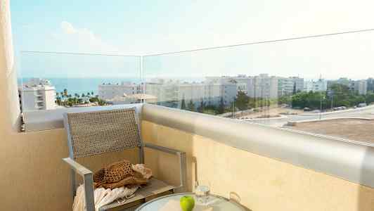 En balkong med utsikt over havet og bygninger, med en stol og et bord med glass, bok og et eple. (AI-generert tekst)