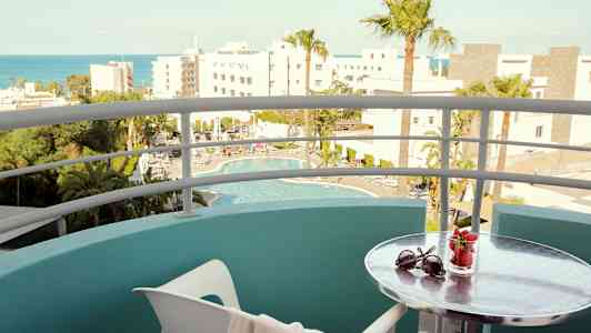 Utsikt fra en balkong mot et basseng og omkringliggende bygninger; et lite bord har solbriller og et glass med jordbær. (AI-generert tekst)