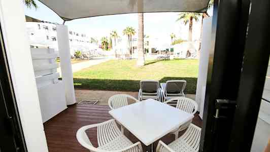 En uteplass med bord og stoler, og utsikt mot grøntområde med solsenger. (AI-generert tekst)