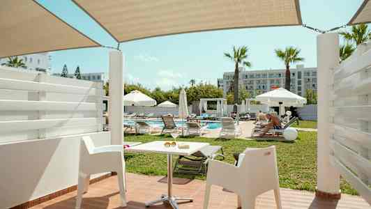 Utsikt fra en terrasse med hvite stoler mot et basseng omgitt av solsenger og parasoller. (AI-generert tekst)