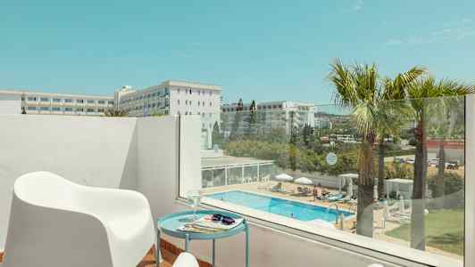 En terrasse med hvite stoler og et bord, utsikt over et basseng og omkringliggende bygninger. (AI-generert tekst)