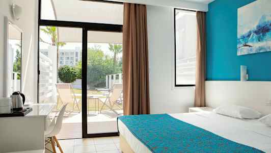 Moderne hotellrom med seng med blått sengeteppe, vindu som åpnes mot terrasse med stoler. (AI-generert tekst)