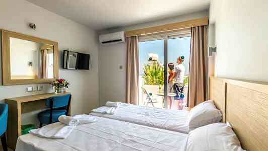 Hotellrom med to enkeltsenger, skrivebord og speil, samt en balkong hvor to personer står. (AI-generert tekst)