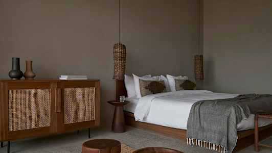 Et soverom med en stor seng, trebord og en kommode med rottingstil. (AI-generert tekst)