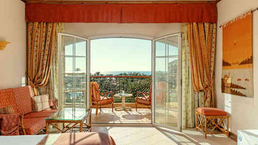 Et hotellrom med åpen balkongdør, sittemøbler og utsikt mot havet. (AI-generert tekst)