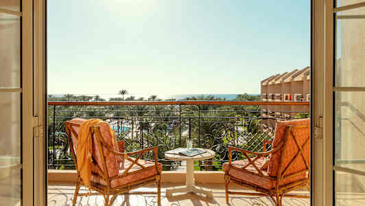 To stoler og et bord står på en balkong med utsikt over palmer og havet. (AI-generert tekst)