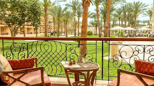 En balkong med stoler, et bord med et glass og solbriller, og utsikt over en hage med palmer. (AI-generert tekst)