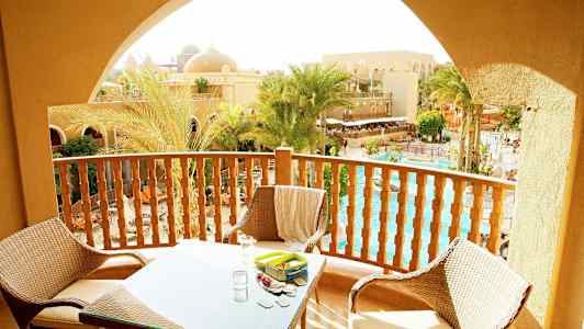 Balkong med møbler med utsikt over palmer, basseng og sandfargede bygninger. (AI-generert tekst)