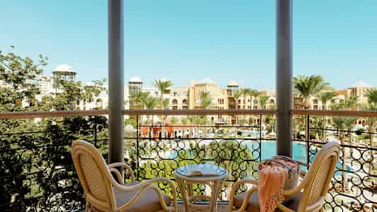 Utsikt fra en balkong med to rottingstoler og et bord. Bak rekkverket ses palmer, et basseng og bygninger med fargerike fasader. (AI-generert tekst)