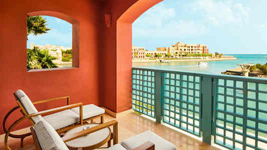 Utsikt fra en balkong med sittemøbler, over en kystlinje og bygninger. (AI-generert tekst)