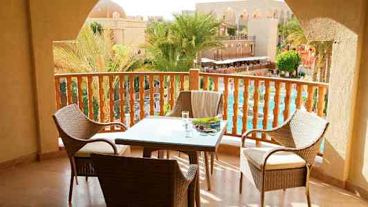 Utsikt fra en balkong med bord og stoler samt utsikt over et basseng og palmer ved en arkitektonisk bygning. (AI-generert tekst)