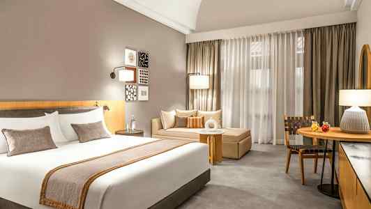 Bilde av et hotellrom med en stor seng, en sofa og et bord foran gardiner. (AI-generert tekst)