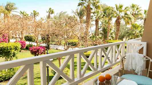 Utsikt fra en balkong med utestoler, bord og appelsiner, med en hage og palmer i bakgrunnen. (AI-generert tekst)