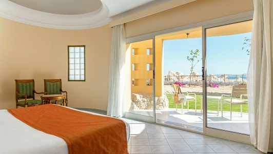 Rom med seng og sengetøy i oransje, to stoler med et bord og utsikt til en terrasse med grøntarealer og solsenger. (AI-generert tekst)