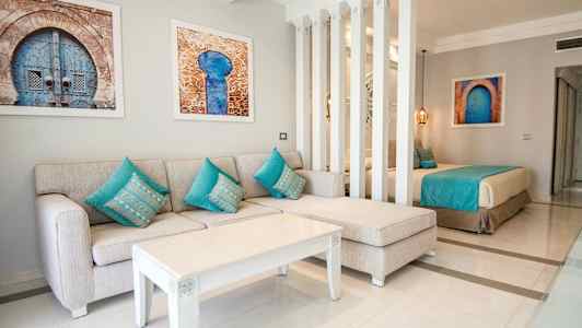 Et oppholdsrom med en beige sofa og turkise puter, et hvitt salongbord og kunstverk på veggen, delvis adskilt med søyler fra en soveområde med en seng. (AI-generert tekst)