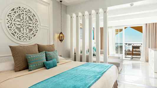 Et hotellrom med lyse vegger, en dekorert seng med blå og beige sengetøy, samt en balkong med havutsikt. (AI-generert tekst)