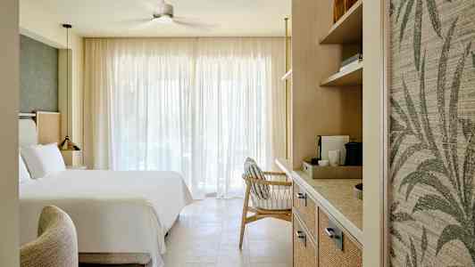 Et hotellrom med en stor seng, en lenestol og hyller med kaffemaskin, opplyst av naturlig lys gjennom store gardiner. (AI-generert tekst)