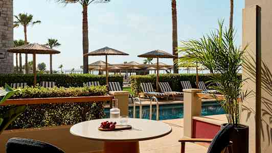 Uteplass med bord, stoler, utsikt mot basseng og solsenger under parasoller. (AI-generert tekst)