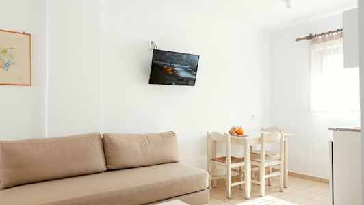 Et lyst rom med beige sofa, veggmontert TV, hvitt spisebord med stoler og et vindu med lyse gardiner. (AI-generert tekst)