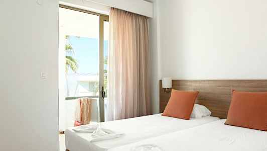 Et hotellrom med to enkeltsenger, oransje puter og en glassdør som fører til en balkong. (AI-generert tekst)