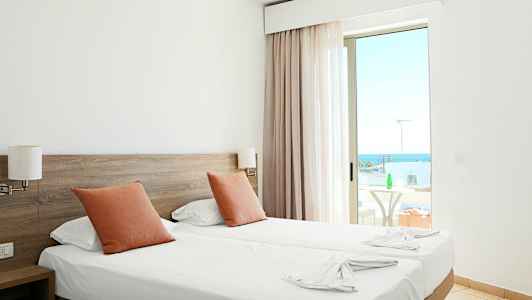 Hotellrom med to enkeltsenger, oransje puter og en balkong med havutsikt. (AI-generert tekst)