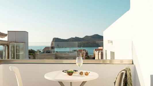 Utsikt fra en terrasse med et bord, en stol og et vinglass samt snacks, bak sees havet og fjell. (AI-generert tekst)