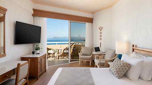 Et hotellrom med en seng, sittegruppe og en balkong med utsikt over havet. (AI-generert tekst)