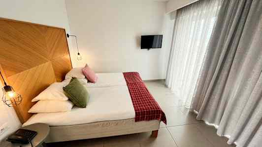 Bilde av et hotellrom med en dobbeltseng, en trepanel som sengegavel, røde og grønne pynteputer, hengende lamper, en liten bordstelefon og gardiner. En TV henger på veggen. (AI-generert tekst)