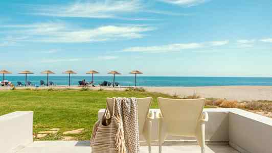 En strand med solsenger og parasoller med havet i bakgrunnen, en uteplass med stoler i forgrunnen. (AI-generert tekst)